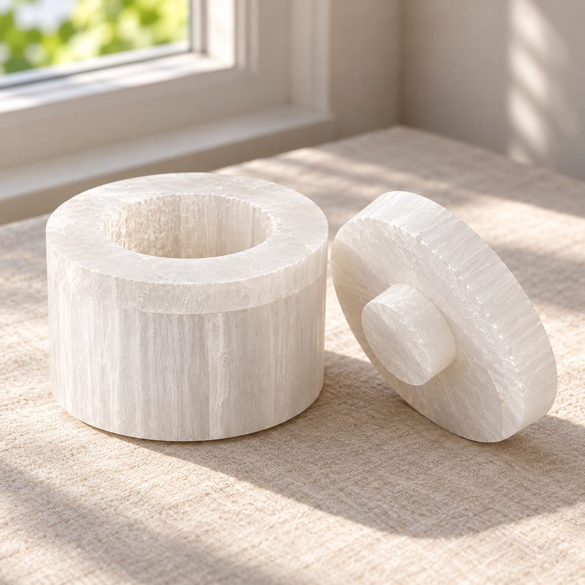 Boîte cylindrique en sélénite ouverte posée près d'une fenêtre — écrin purificateur artisanal fait main au Maroc, pierre naturelle blanche | Pure Selenite